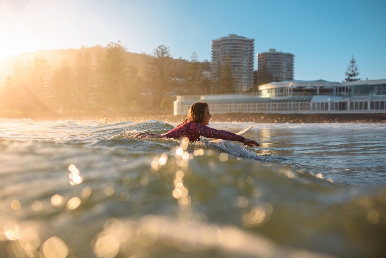 Surface level side view with lens flare of smiling woman in early 20s paddling into shore at Burleigh Heads.